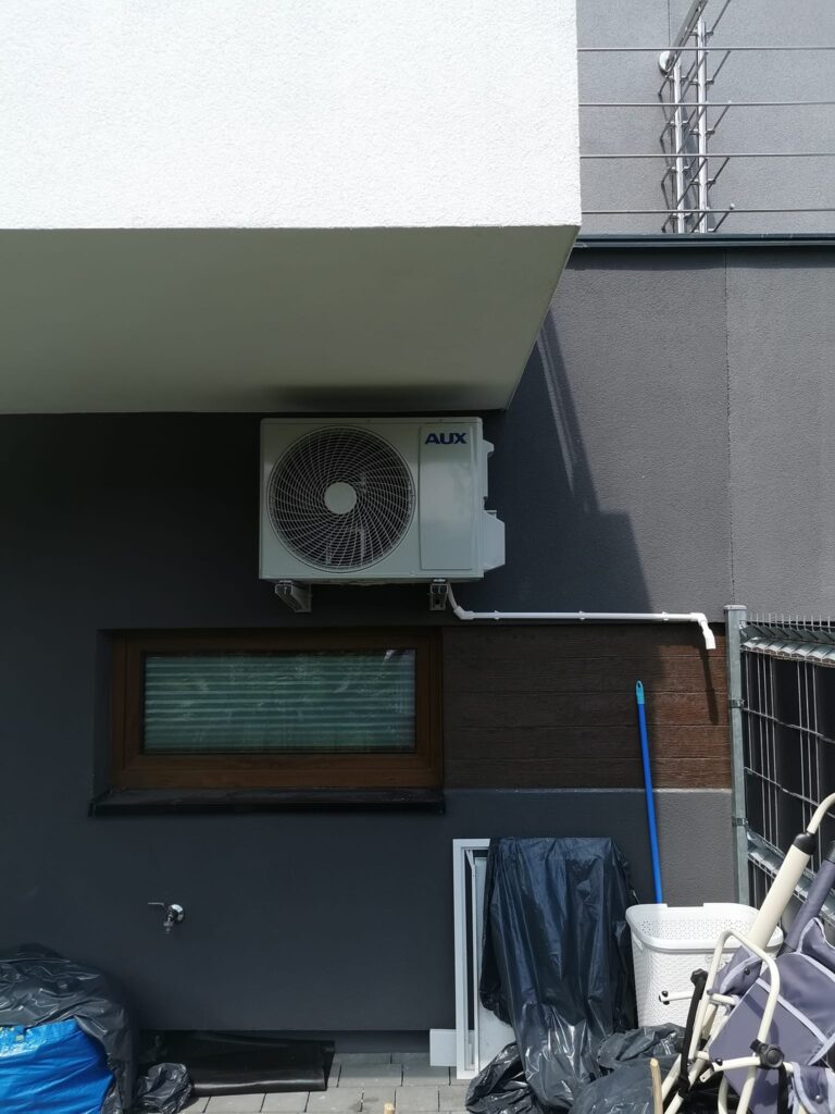 zdjęcie przedstawia jednostkę zewnętrzną klimatyzacji marki AUX (agregat) zamontowany na ścianie domu jednorodzinnego.